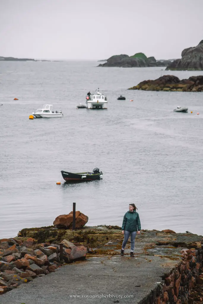 woman stands at the edge of a pier on the isle of mull in scotland with boats in the backgrund